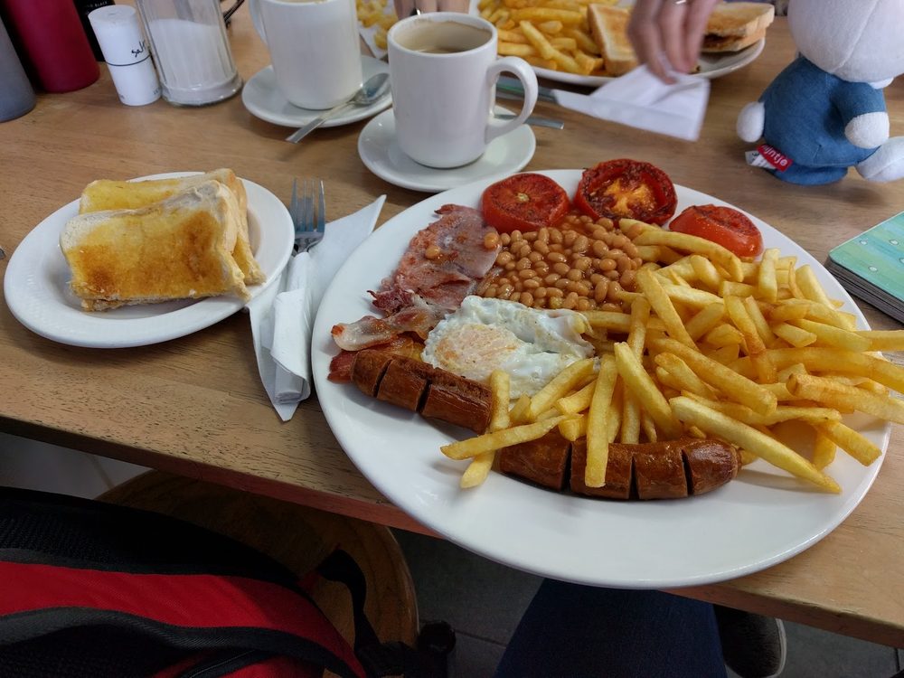 A loaded breakfast plate on a busy shared table with mugs and cutlery around it